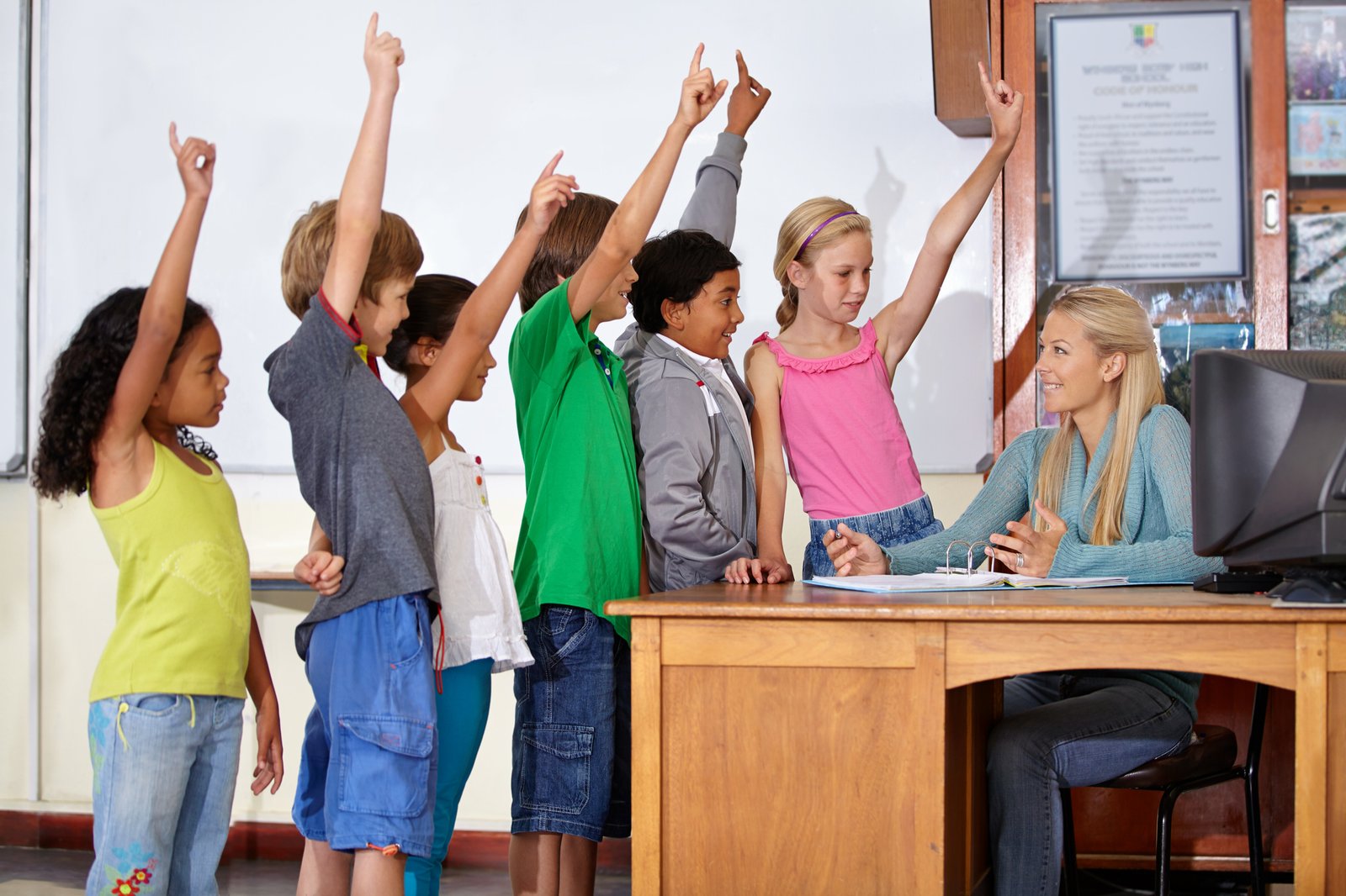 Teaching is her dream. A teacher sitting at her desk talking to her students who all have their hands up.